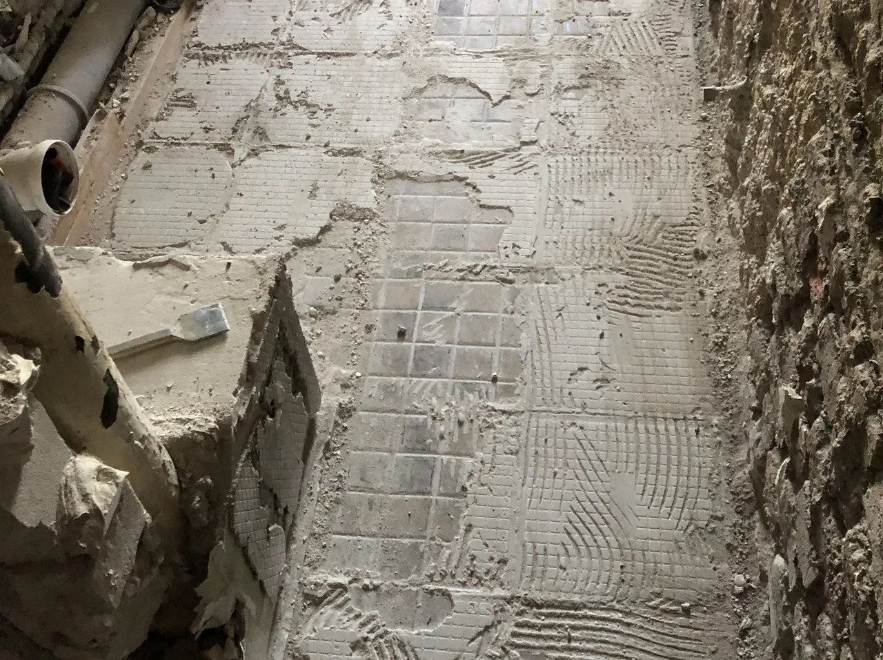Abgeschlagener Fliesenboden in einem Altbau: Reste von Mosaikfliesen im Kleberbett, Zahnspuren des Klebers im Untergrund und Abflussrohre am Wandfuß