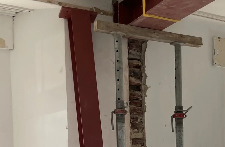 Durchbruch in einer tragenden Altbauwand im Rohbau: ein roter Stahlträger unter der Decke, ein roter Stahlstiel und zwei Baustützen vor einem freigelegten Ziegelstreifen