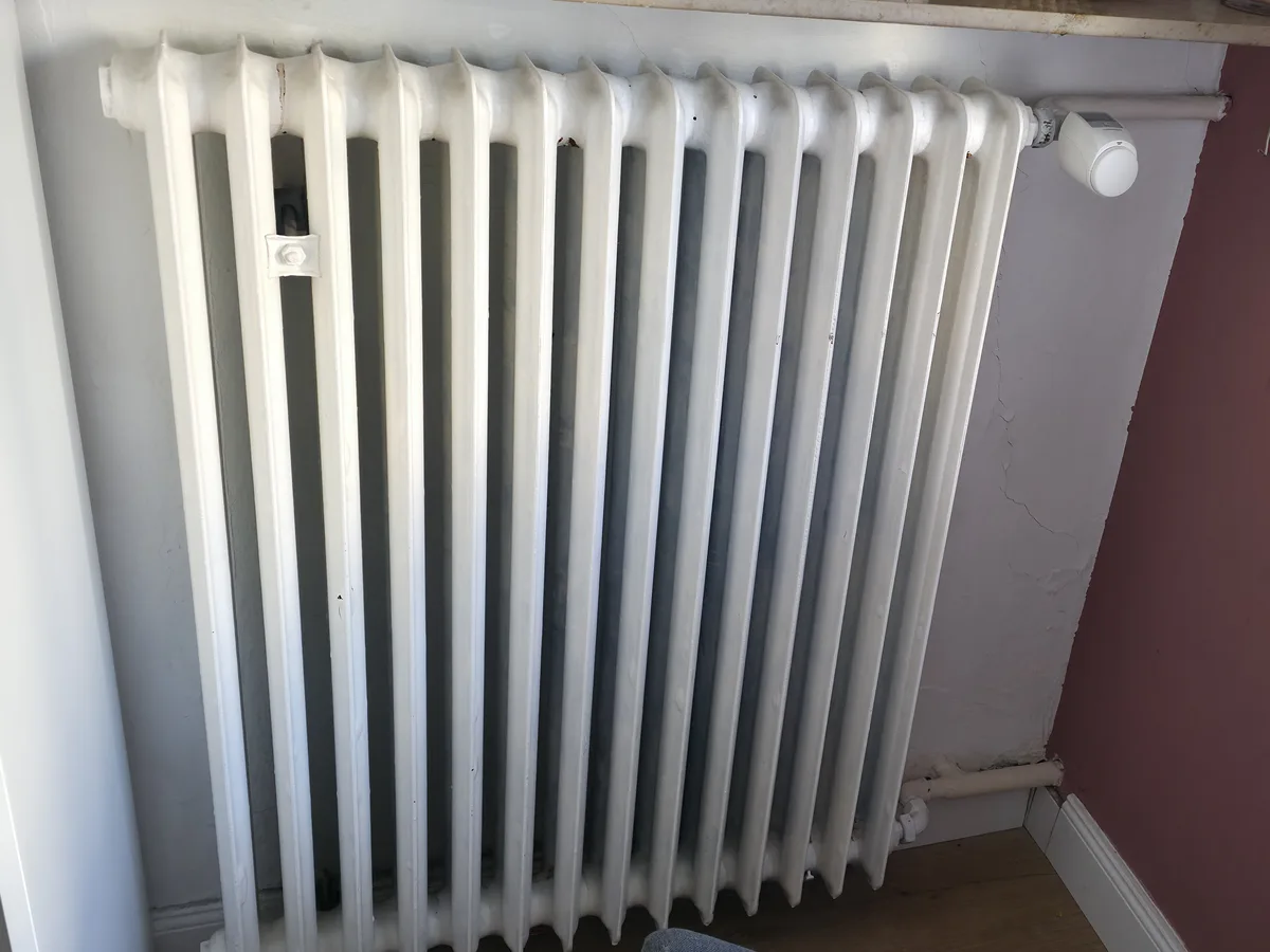 Weißer Altbau-Gussradiator mit vielen schmalen Rippen und Thermostatkopf unter einer Fensterbank, daneben der Wandanschluss mit den Anschlussleitungen