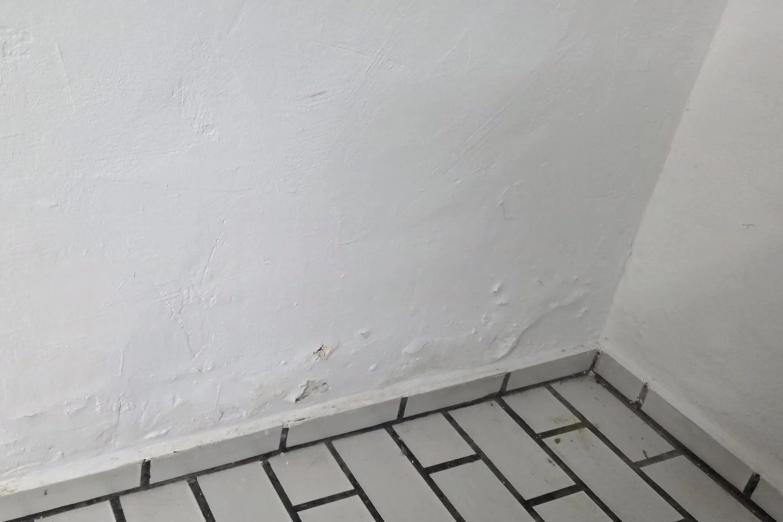 Weiß gestrichene Kellerwand über einem Fliesenboden: im Sockelbereich Farbblasen, Abplatzungen und aufgeplatzte Stellen im Anstrich