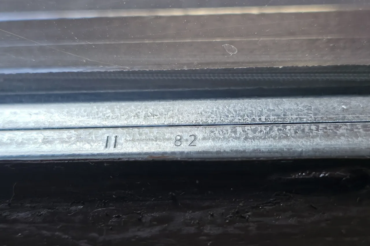 Nahaufnahme des Aluminium-Abstandhalters im Scheibenzwischenraum einer Haustür-Verglasung mit eingestanzter Jahreszahl II 82 über dunklem Holzrahmen