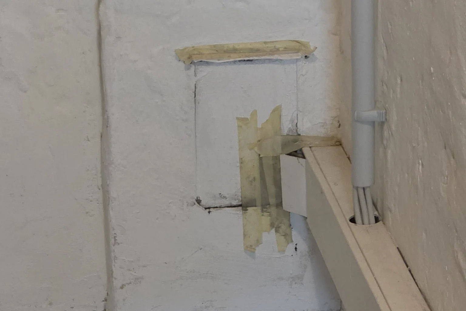 Weiß gestrichene Kellerwand mit einer rechteckigen Reinigungsluke, die mit Klebeband verschlossen ist, daneben eine Kabelleiste