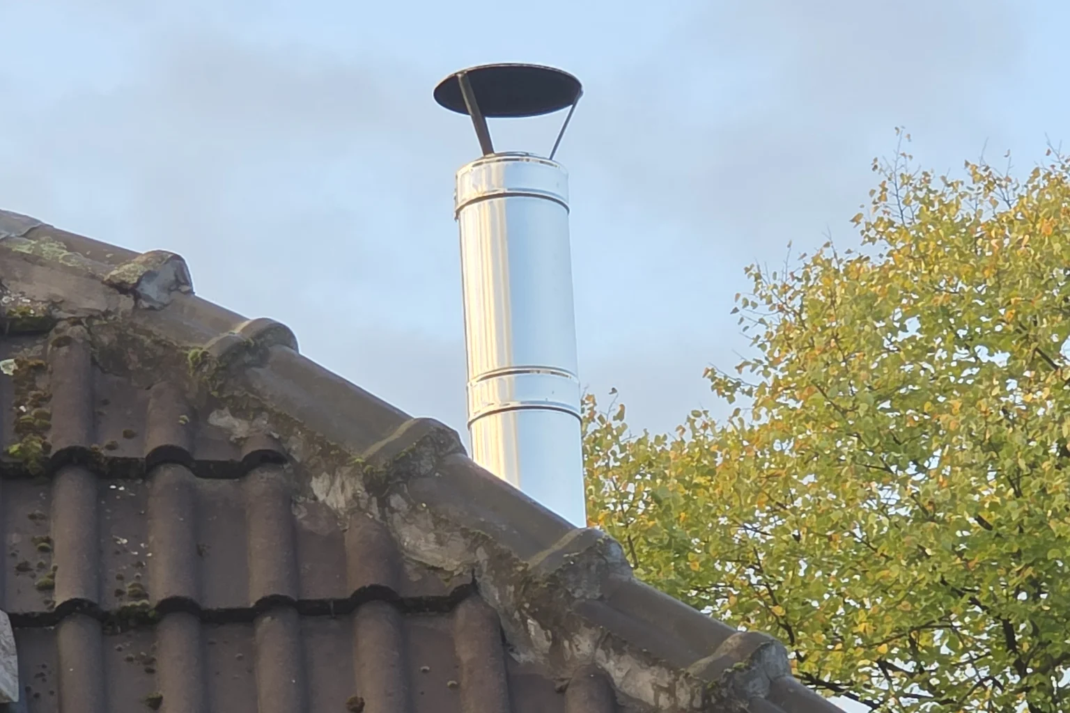 Edelstahl-Abgasrohr mit Regenhaube über dem Ziegeldach, am First bemooste Ziegel und bröckelnder Mörtel