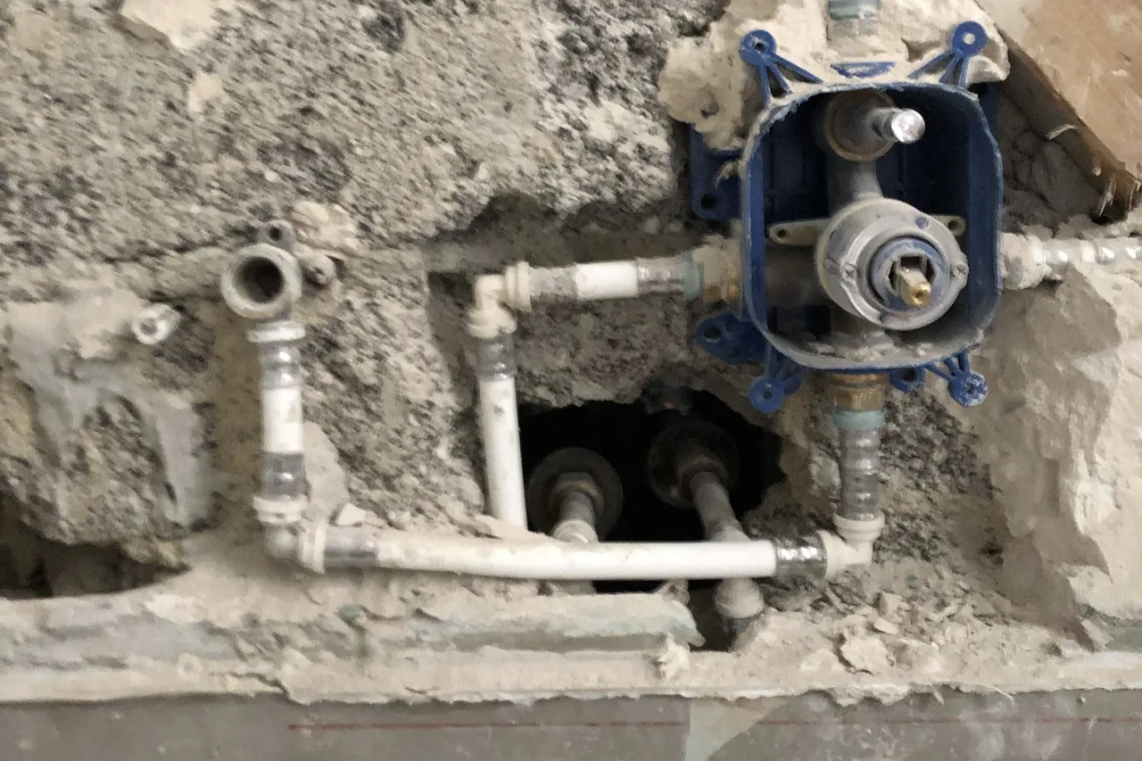 Aufgestemmte Wand im Bad: weiße Verbundrohre mit Pressfittings um einen blauen Unterputz-Einbaukörper, darunter ein dunkler Hohlraum mit weiteren Rohren